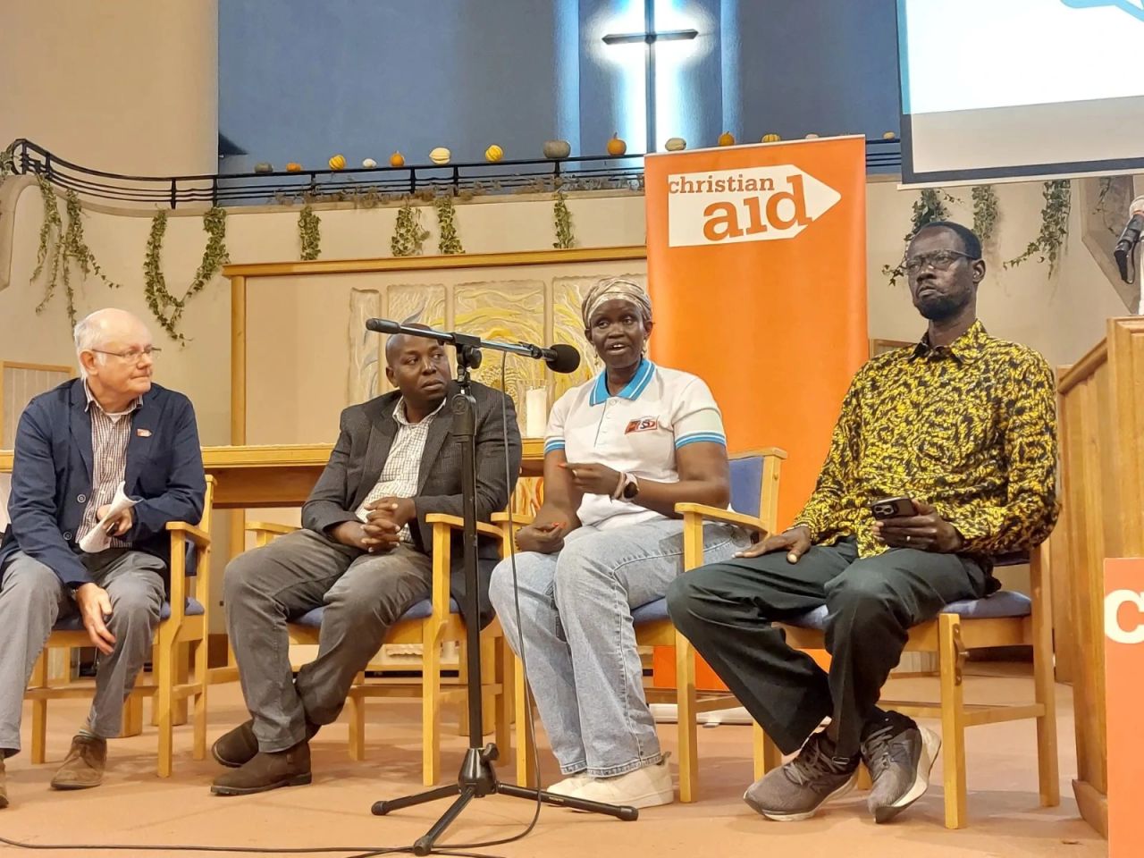 Angelina Jial, Founder and Executive Director of HRSS, speaks into a microphone at a panel discussion in a church hall, seated with three other panellists in front of a Christian Aid banner.