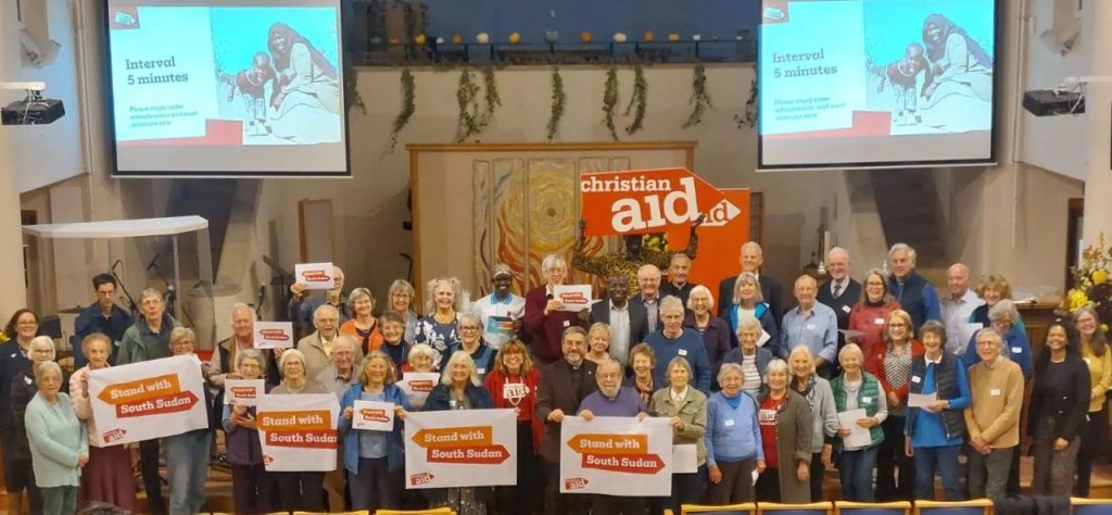 Large group of supporters standing together in a church hall, holding “Stand with South Sudan” signs in front of a Christian Aid banner.
