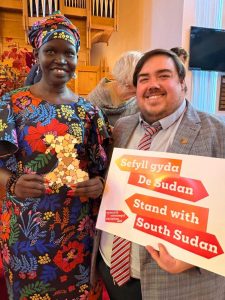 Angelina Jial holds a map of Wales made of small wooden hearts, next to a man holding a bilingual Welsh and English “Stand with South Sudan” sign.